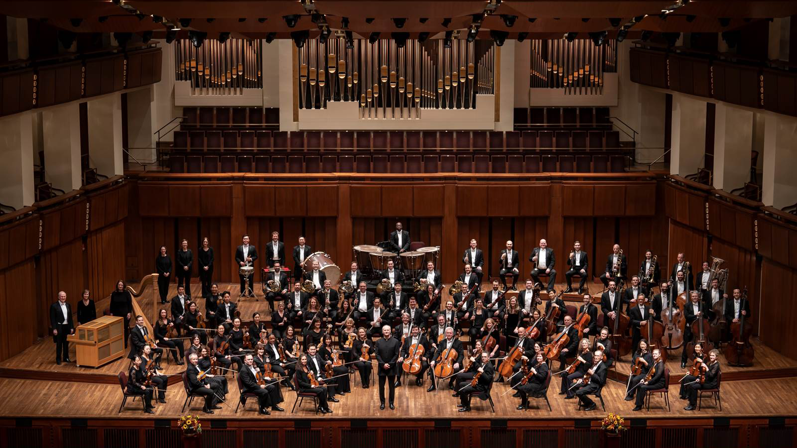 A photograph of the entire National Symphony Orchestra with their instruments and Music Director, Gianandrea Noseda, on the Concert Hall stage with the massive pipe organ behind it. The photo is taken from the balcony level. Everyone is looking up at the camera. Maestro Noseda is standing in front and his hands are clasped in front of him.