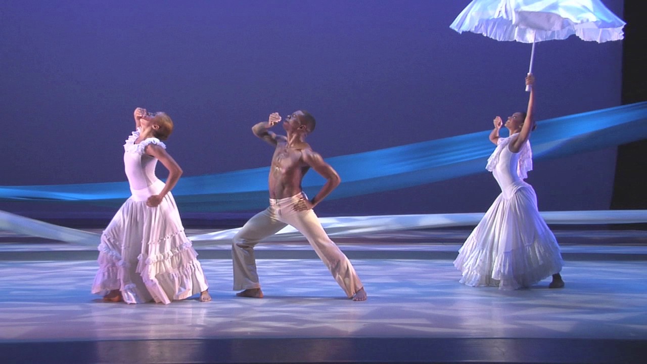 Three barefoot dancers (two women and one man) move with fluidity in a scene that recreates a baptism ritual. The women wear long sleeveless white dresses with flowy, ruffled skirts while the man wears linen pants. One woman holds a flowy white umbrella in the air.