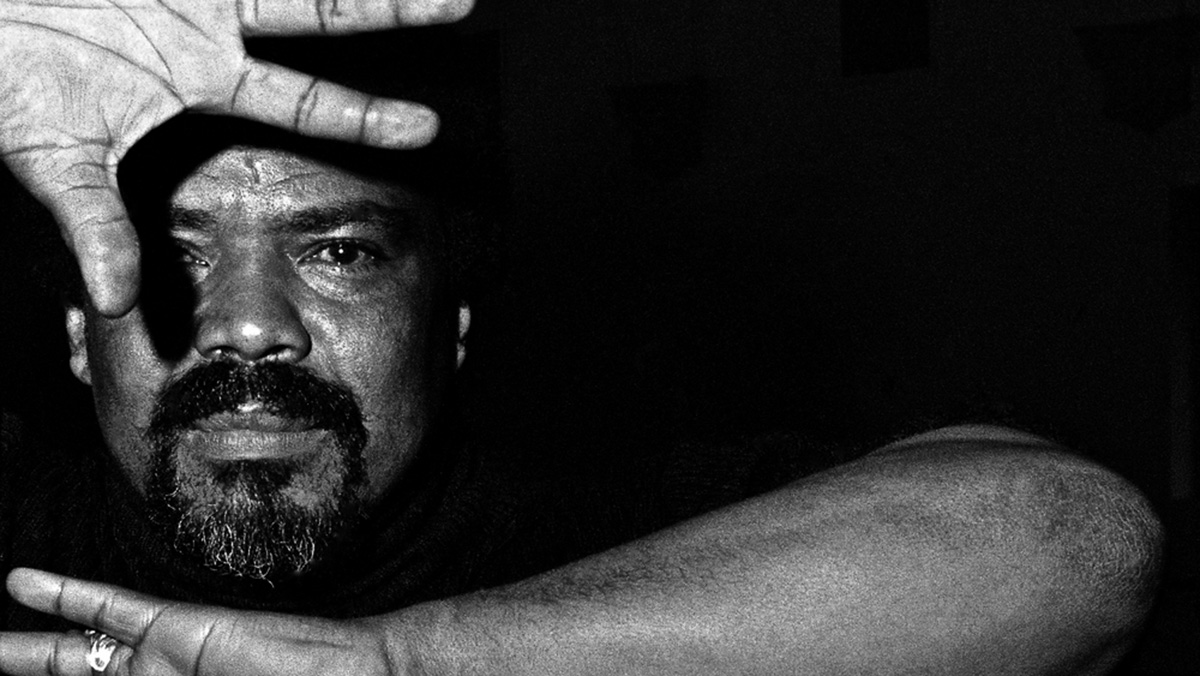 A black and white headshot of dancer and choreographer Alvin Ailey. He holds his hands in front of his forehead and neck with the palms facing towards the camera. The lighting throws a sharp shadow over his face.