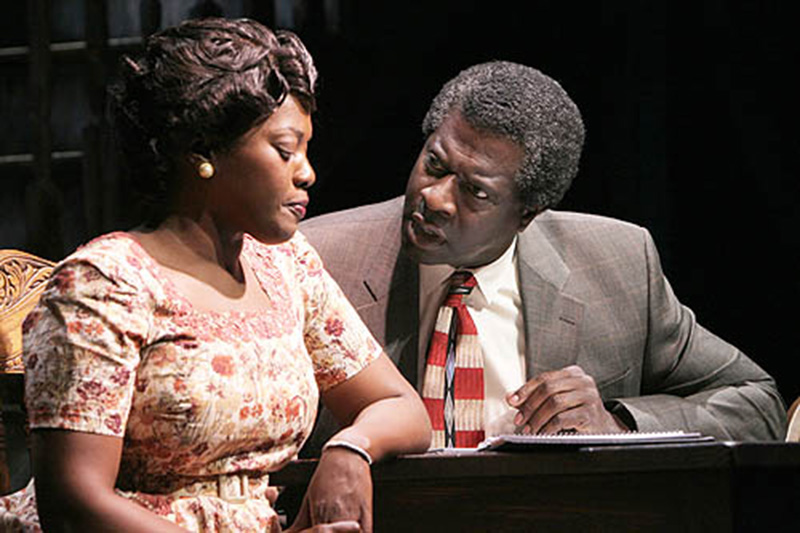 In a staged scene, a man with graying hair talks to a woman sitting next to him at a table. He looks at her with his arm resting on a notebook while her eyes are cast downward. He wears a gray jacket and a white dress shirt with a red-and-cream striped tie. She wears a beige dress with a burgundy-and-green floral pattern and pearl earrings.