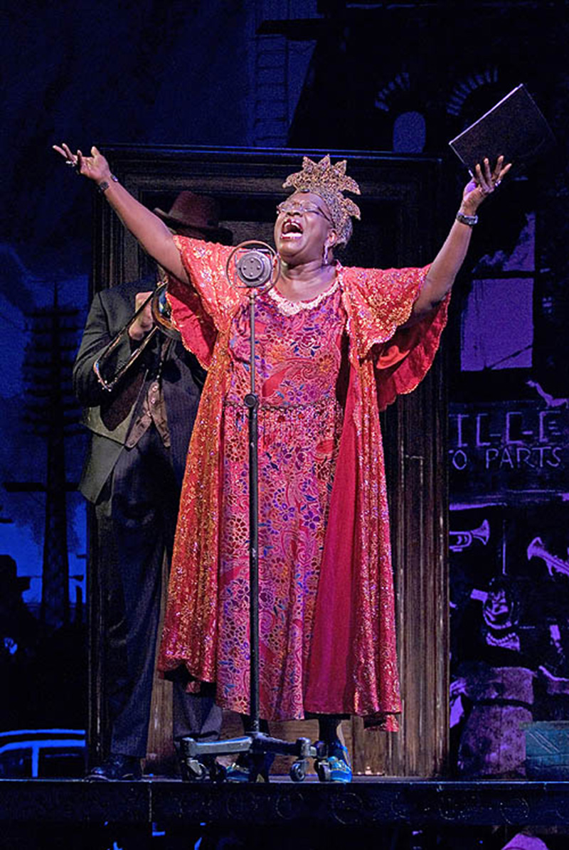 In a staged scene, a woman sings at an old-fashioned microphone stand with her arms raised mid-performance. She wears a long dress with draped elbow-length sleeves in red, gold, and purple. An ornate headband/wrap rests on her head, along with matching bracelets on both wrists. Behind her is a man in a dark suit and hat playing a trombone.