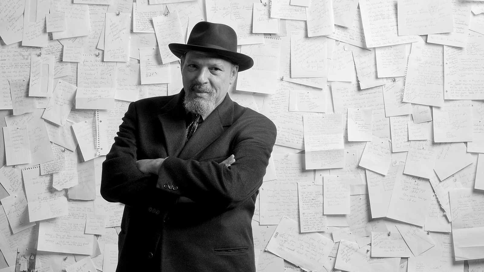 A black and white photo of playwright August Wilson wearing a dark jacket with a matching dark tie and hat. With his arms crossed, he stands in front of a wall on which many pieces of handwriting-covered notebook and notepad paper are tacked.
