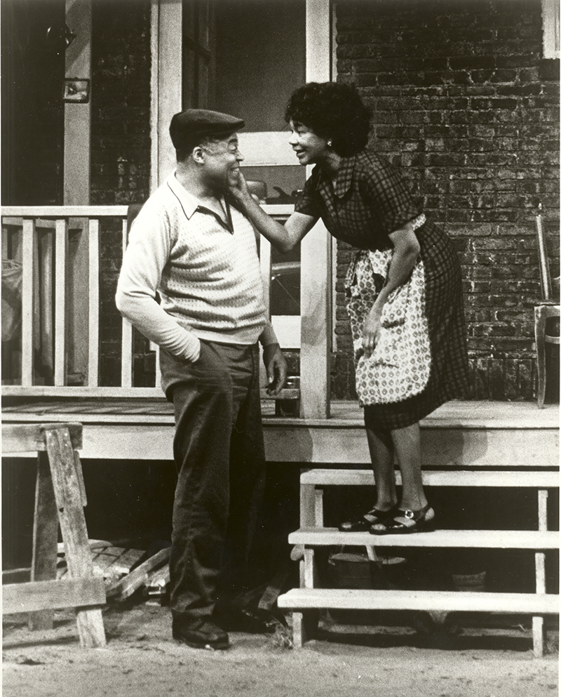 A vintage photo of a staged set where married characters Troy and Rose smile at each other in front of their home. Standing on patio steps, Rose touches her husband&rsquo;s face with love. She wears a dark plaid dress with an apron and sandals while he wears a patterned long sleeve shirt with dark pants and a matching news cap.