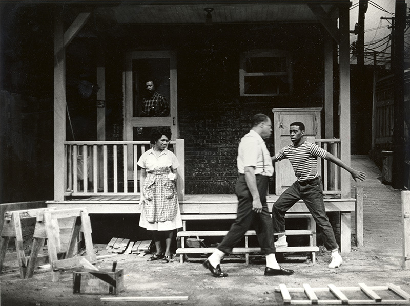 In a black-and-white photo of a stage set to look like a house and its front yard, a woman watches as her husband walks by her and their son. The son&rsquo;s arms are spread out mid-action. Inside the house&rsquo;s screen door, another man can be seen looking out at the scene. 
