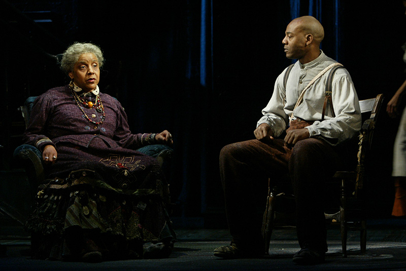 A scene from one of Wilson&rsquo;s staged plays, an elderly woman sits in a dark teal armchair next to a man sitting in a wood chair. The man wears a light-colored shirt with dark brown pants and suspenders, and he looks at the woman. The woman wears a dark purple outfit with multicolored layered necklaces.