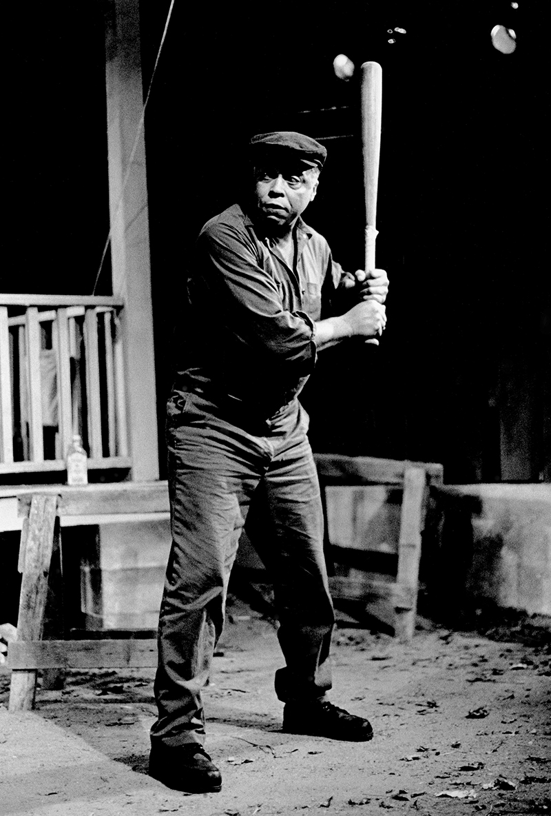 A black and white photo of a man on a stage set to look like a yard in front of a patio. The man holds a baseball bat upright in both hands and looks off to his right.