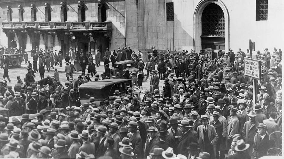 A vintage photo of an overcrowded city street filled with people wearing trench coats, business suits, and business hats. The crowd lingers outside a building.jpg