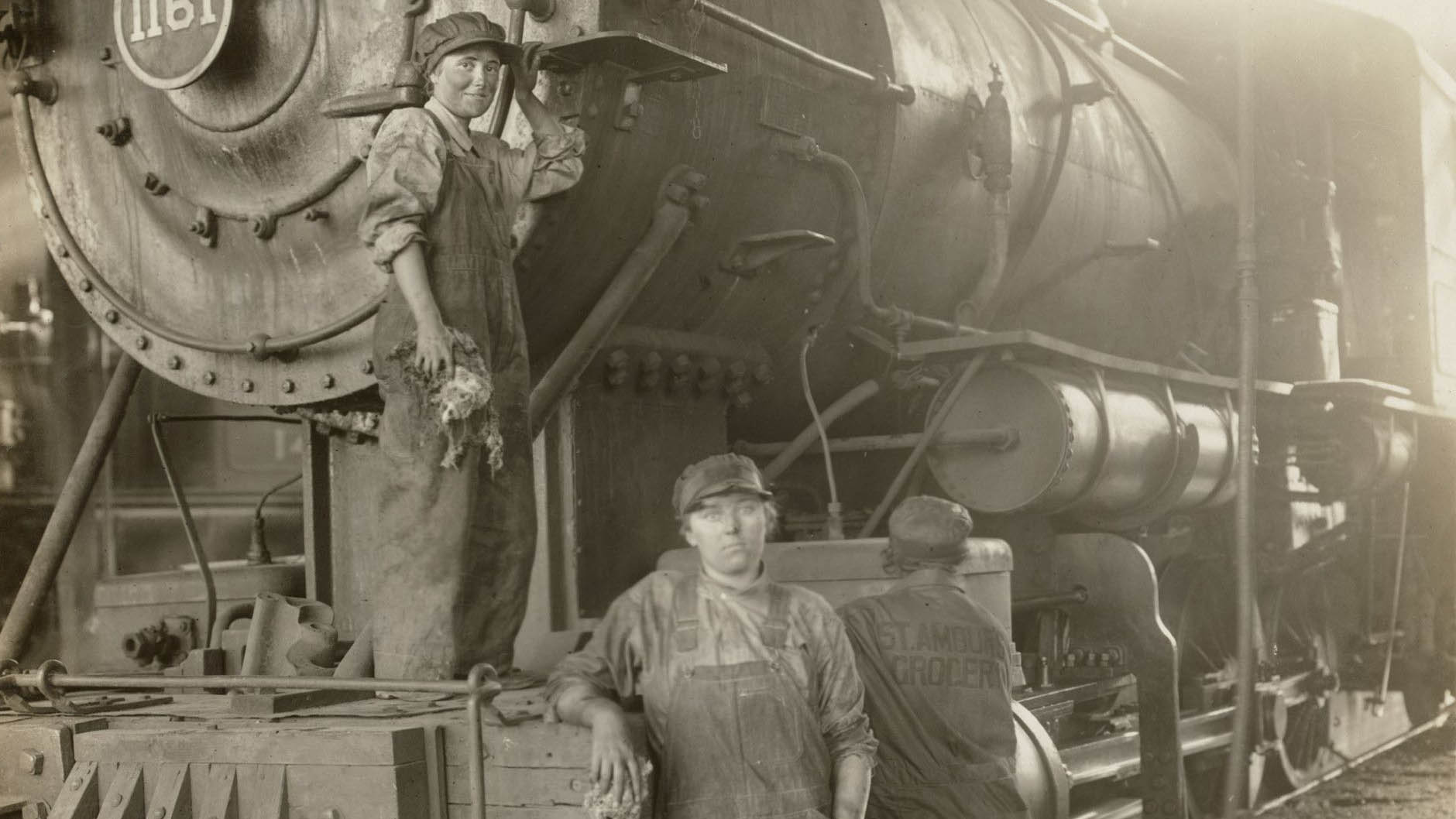 A vintage photo of female workers dressed in overalls, long-sleeved shirts, and railway hats standing beside a train.jpg