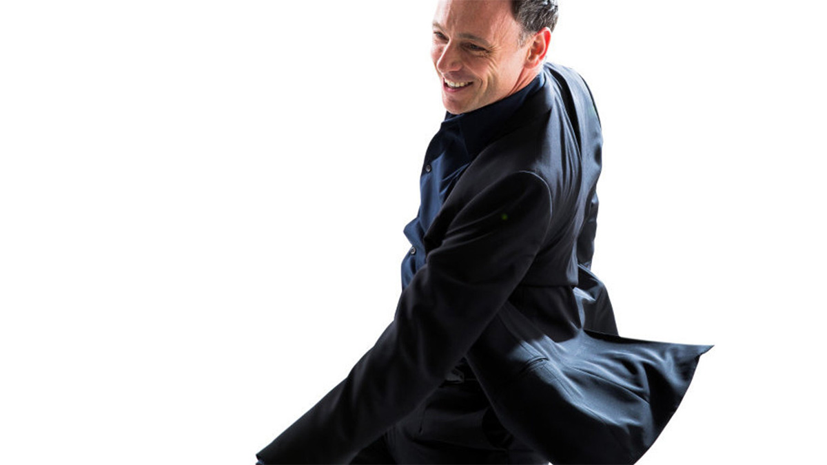 A photo of a smiling man in a dark blue jacket is captured as he is spinning, the back of his jacket billowing upward.