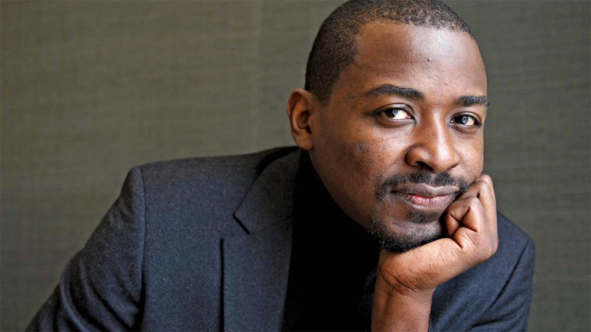 A headshot of a man resting his chin in his left hand. He wears a textured, dark blue blazer and a dark shirt.