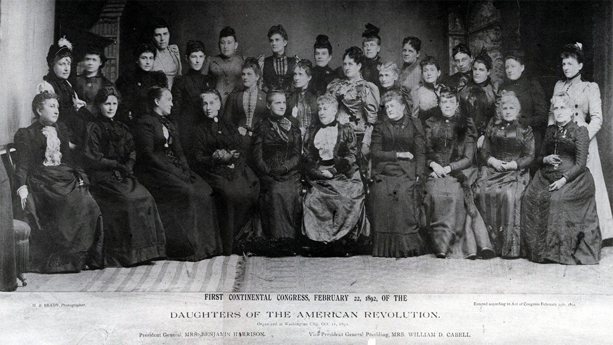 A black and white photo taken in 1892 of the Daughters of the American Revolution (or D.A.R.). Three rows of white women pose in period dresses, updo hairstyles, and the occasional hair accessory; the front row of women sit while the back two rows stand.