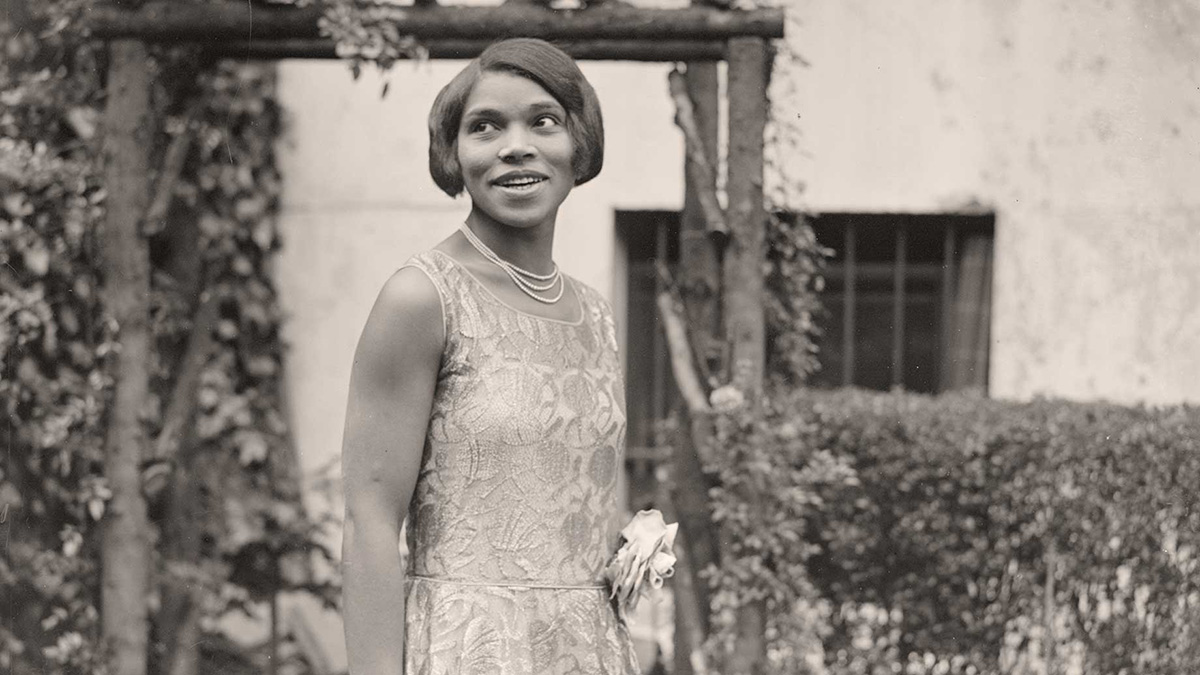 A black and white photo of a young Marian Anderson. Set outside of a building and in front of a wood arch, Marian wears a sleeveless dress with an intricate pattern overlay and a big flower accessory on the waist, as well as a three-strand necklace.