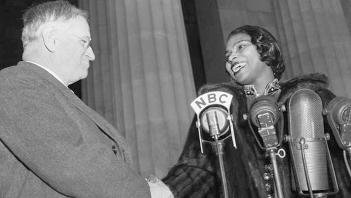 A black and white photo from 1939 in which Harold Ickes, Secretary of the Interior, shakes hands with singer Marian Anderson. Ickes, wearing a dark business jacket and glasses, is turned towards Anderson, wearing a fur coat while standing in front of four vintage microphones. They are both smiling. Columns from the Lincoln Memorial are visible behind them. 