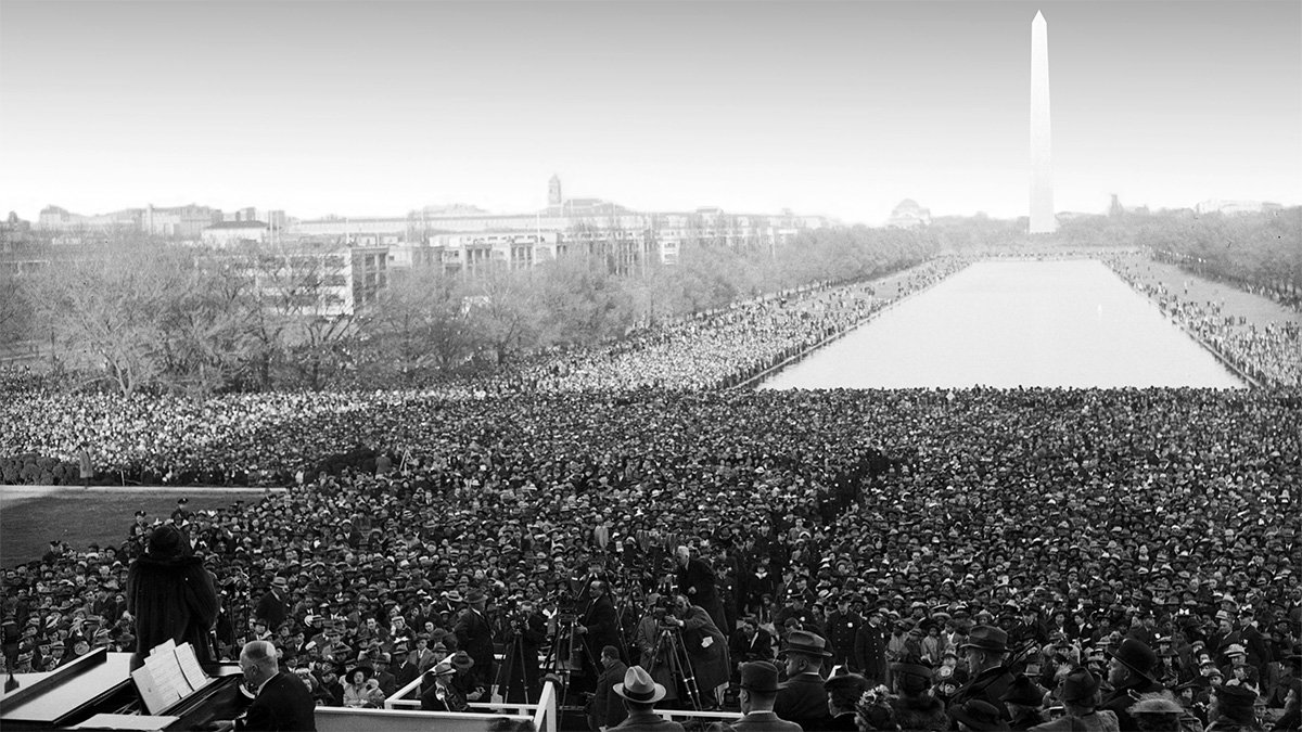 A black and white photo taken in 1939 of Marian Anderson performing at the Lincoln Monument in front of a huge crowd. The back of Marian Anderson is visible in the bottom left corner of the photo; she stands in front of a pianist. There are thousands of people standing next to each from the end of the stage (near the bottom edge of the photo) all the way around the reflecting pool towards the Washington Monument (towards the top right corner of the photo).