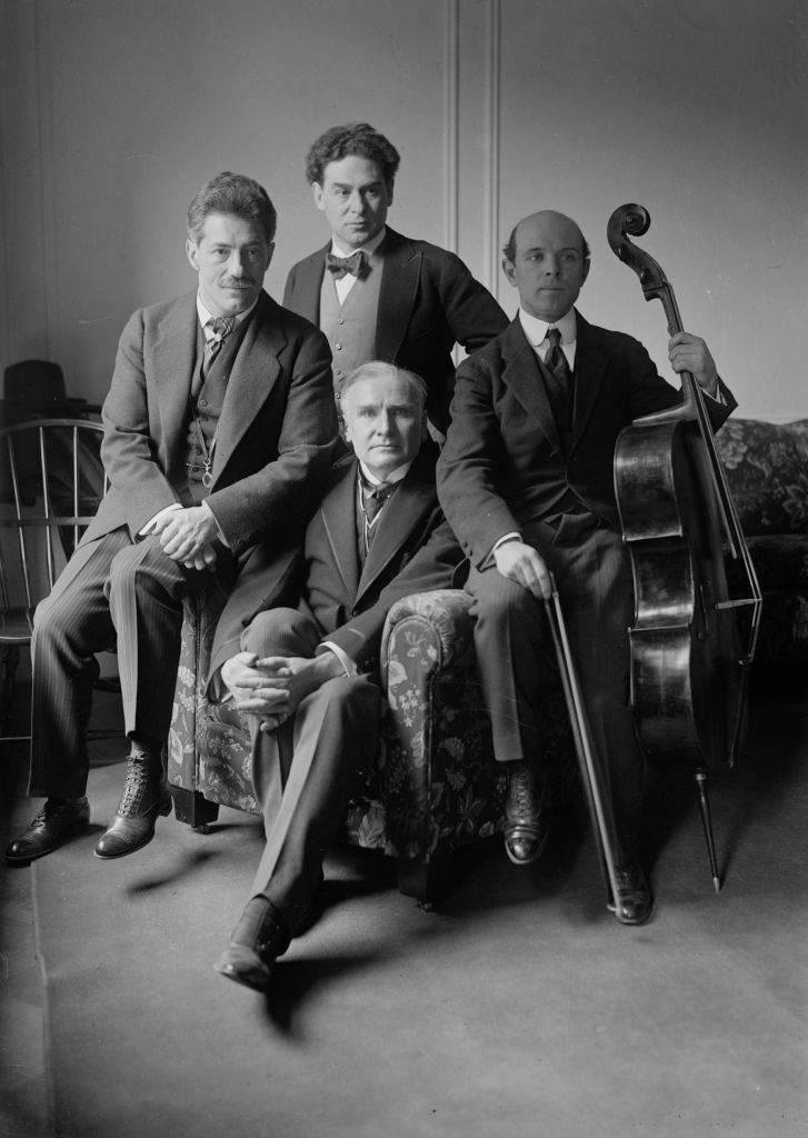 Black and white photo portrait of musicians Fritz Kreisler, Walter Damrosch, Pablo Casals, and Harold Bauer sitting with their instruments.