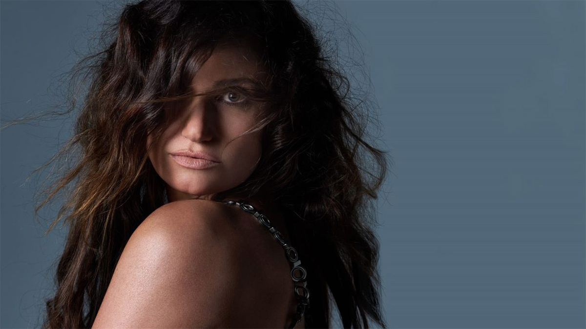 A head shot of singer Idina Menzel in front of a light blue background. Her hair is curly and part of her hair sweeps over her right eye.