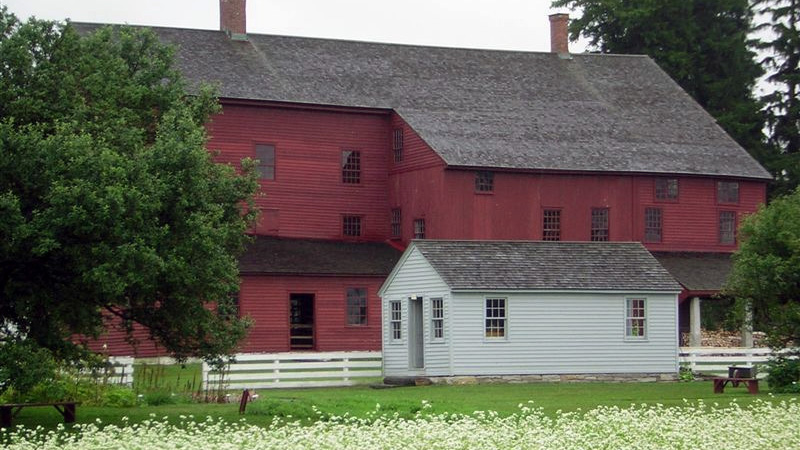 A photograph in which a small white house is set in a green field with white flowers in the foreground and green trees on the photograph&rsquo;s left and right edges. Behind the small house is a white fence and a very large red building.