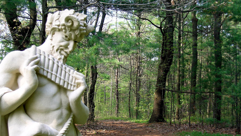A photograph of a mischievous-looking half-man, half-goat statue playing a pan flute in a forest. 