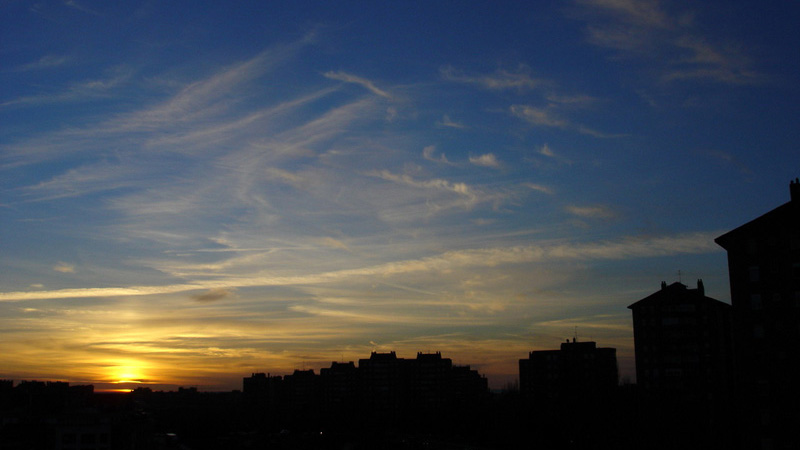 A photograph of the setting sun against a sky with brushstrokes of clouds. The horizon is lined with the silhouettes of building tops.