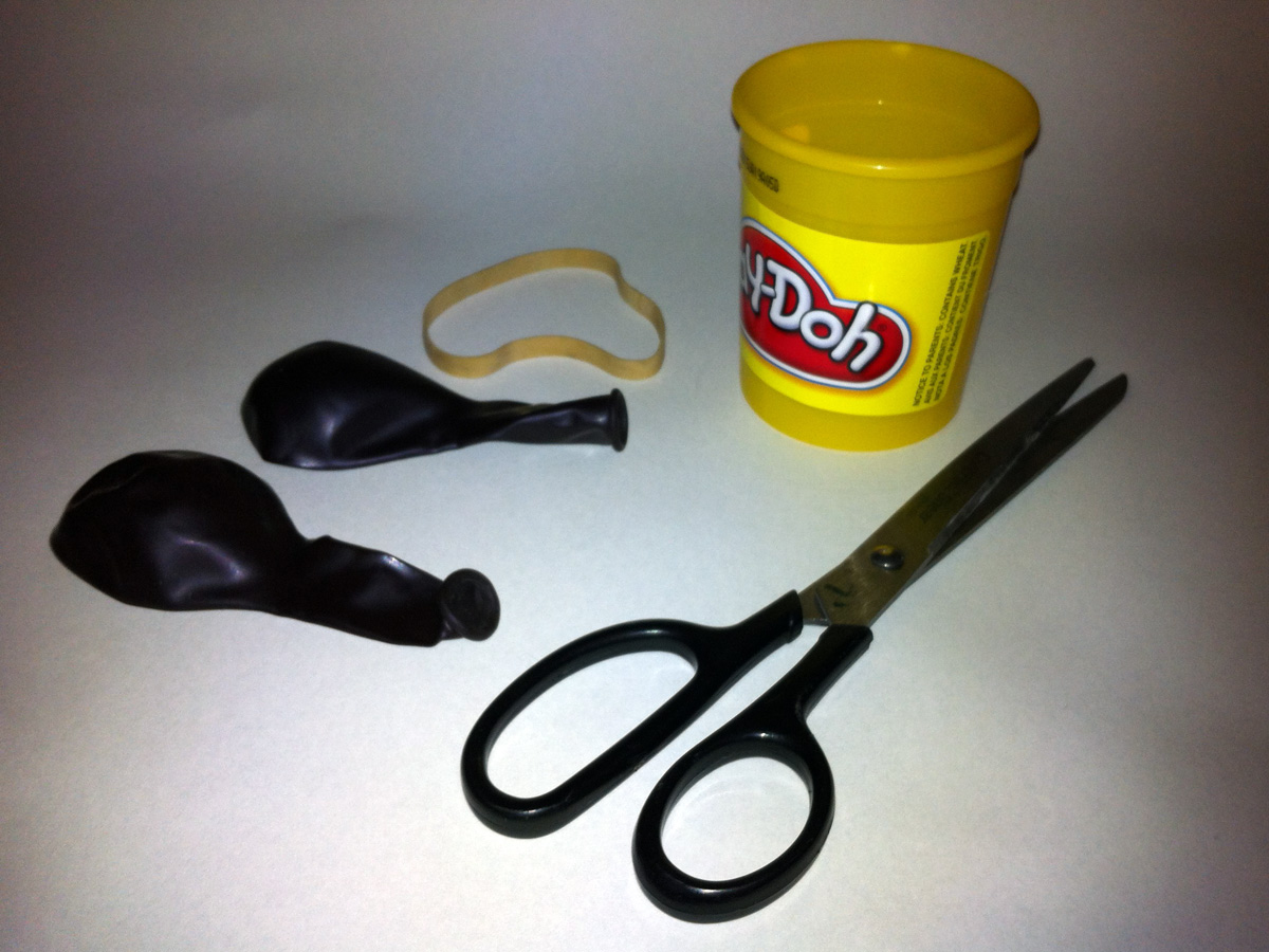 A photo of two balloons, a rubber band, a pair of scissors, and an empty Play-Doh container.