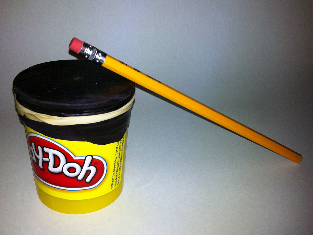 A photo of a completed do-it-yourself drum made with a Play-Doh container, balloons strapped across the open top with rubber bands, and a pencil as the drum stick.