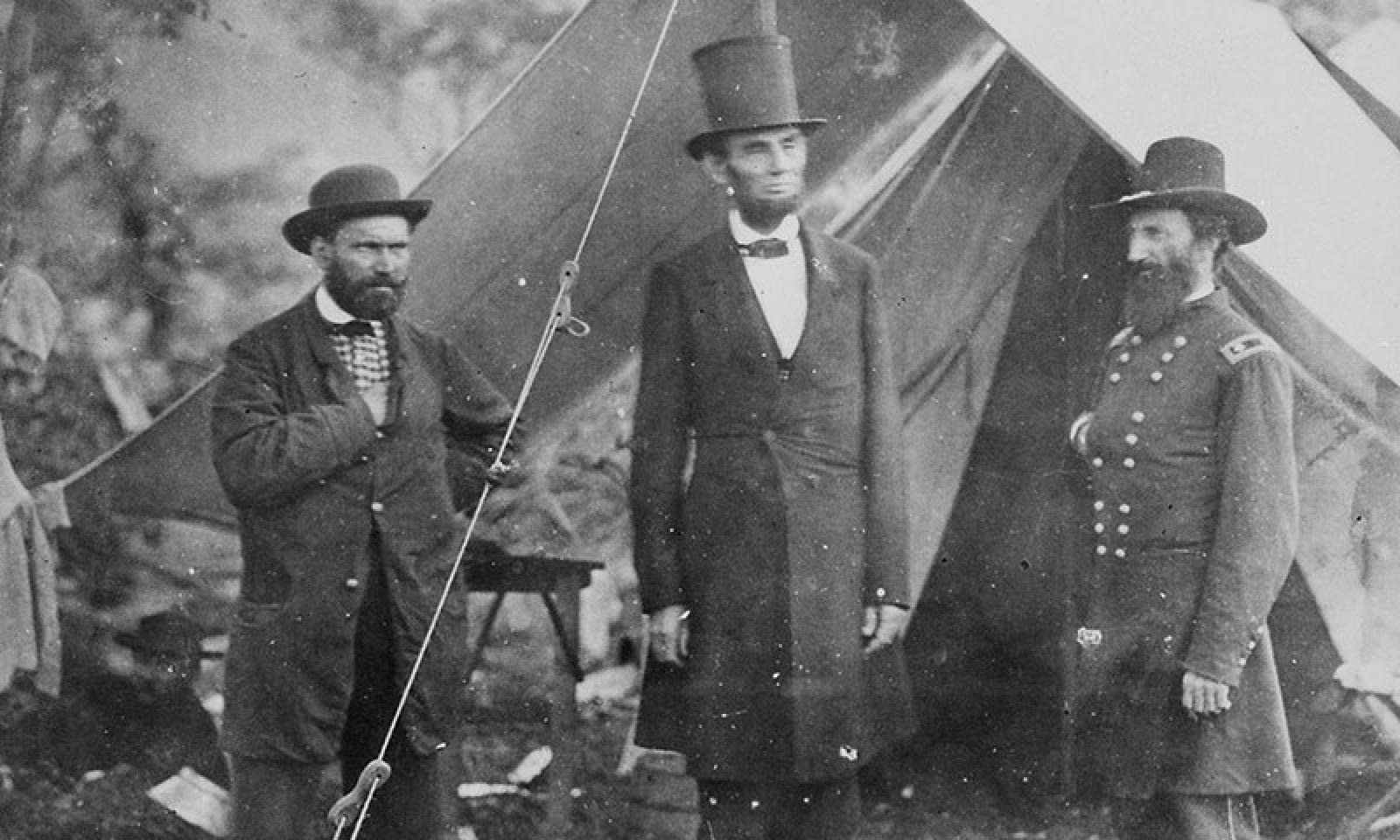 An image of President Abraham Lincoln standing beside two men (one wearing a military uniform) in front of tent. Lincoln wears a long coat and a stovepipe hat.