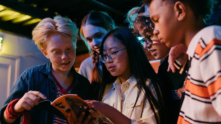 Five diverse teenagers stand close to each other to look at a comic book. 