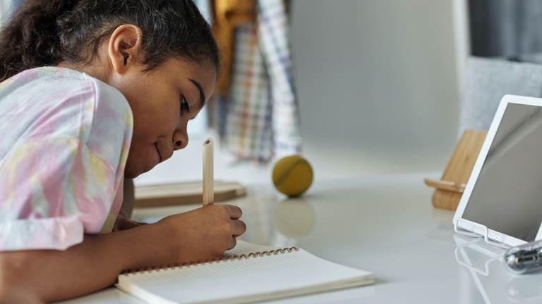 A young Black person writes in a notebook while sitting at a desk.
