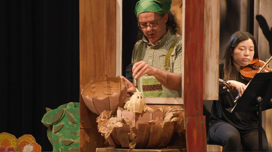 A production photo of Old MacDonald revealing a baby chick puppet inside of a large cardboard-constructed chicken egg. Old MacDonald wears a patchwork-patterned green handkerchief on the head with a green plaid shirt, green-and-brown patchwork overalls, and round eyeglasses. Behind Old MacDonald is one of the violinists from the NSO performing while sitting in front of a music stand.