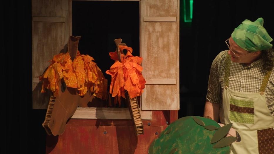 A production photo of Old MacDonald looking at two horse puppets whose heads peek out from horse stall doors. The horse puppets are made from brown cardboard and have orange manes. Old MacDonald wears a patchwork-patterned green handkerchief on the head with a green plaid shirt, green-and-brown patchwork overalls, and round eyeglasses.