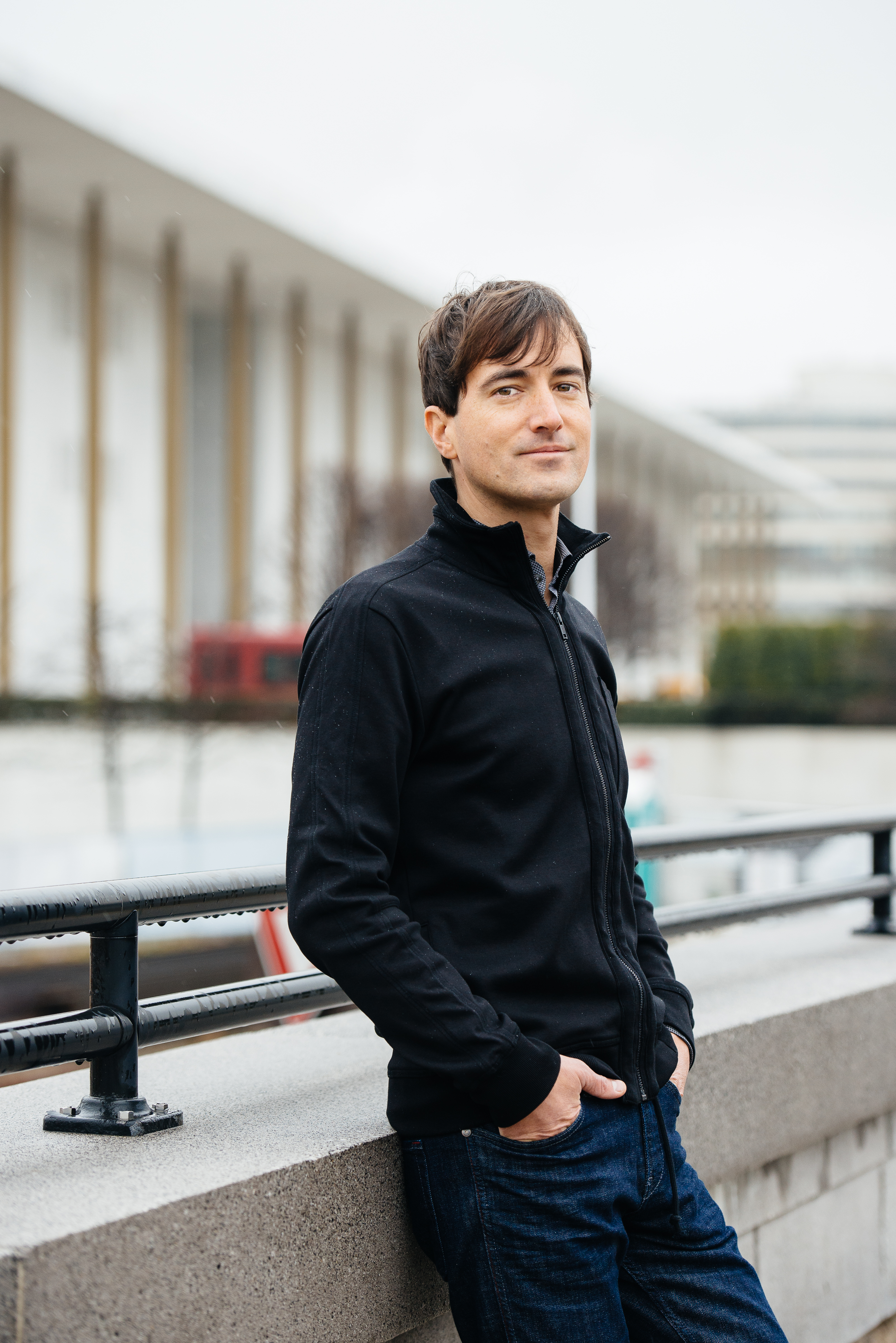 Photo of Mason Bates by Kate Warren. A tall slim white man with short brown hair dressed in jeans and a dark blue sweater is standing with hands in his pockets outside in front of the Kennedy Center.