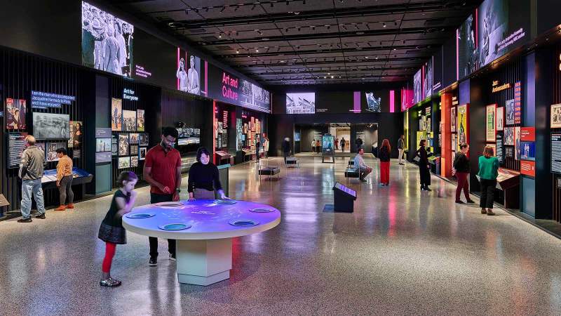 Interior of exhibit with people browsing artwork and an interactive table, surrounded by screens.