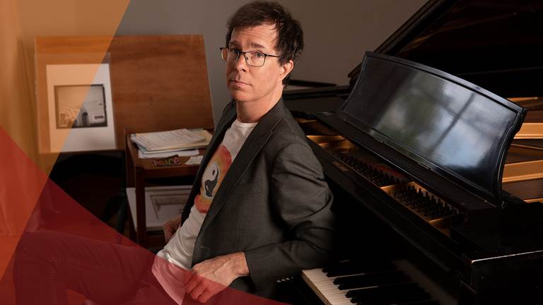 Ben Folds leaning back against piano in t-shirt and blazer