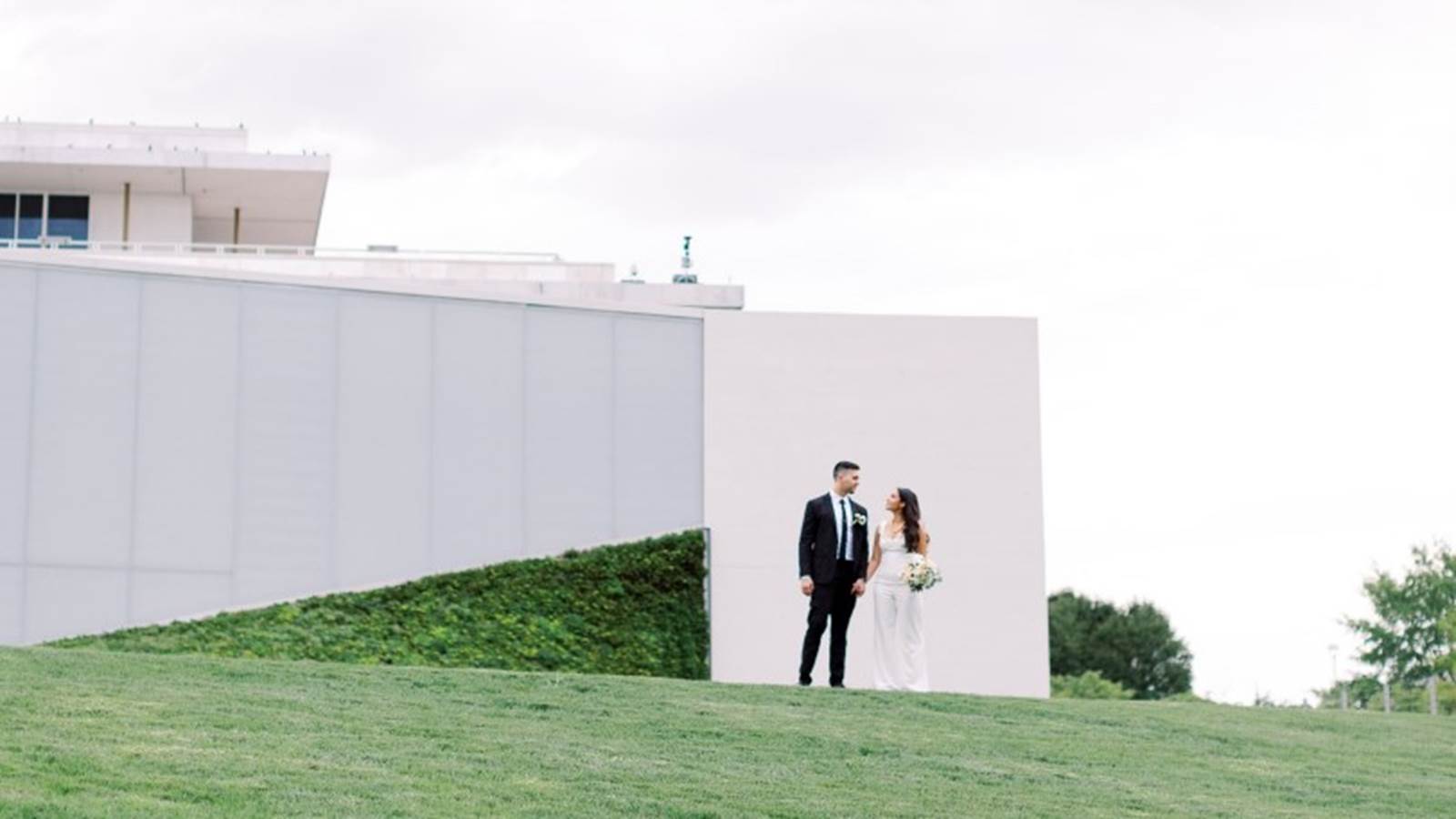 Bride and groom stand on a field of grass in front of the Skylight Pavilion at the REACH