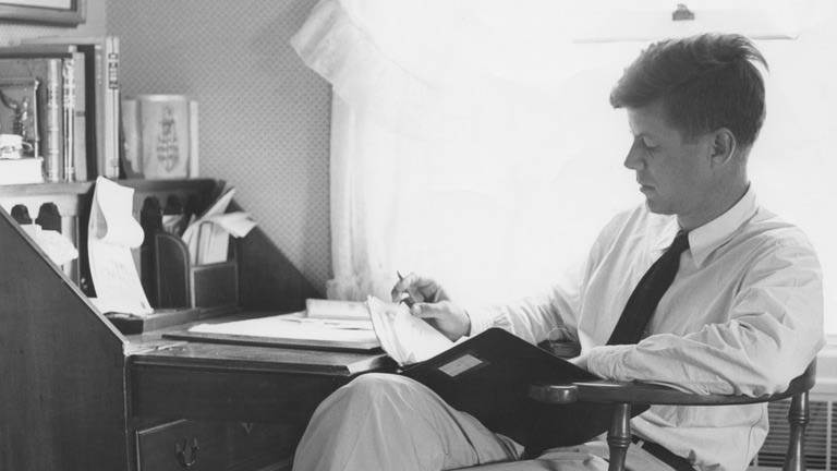 John F. Kennedy reading at a writing desk