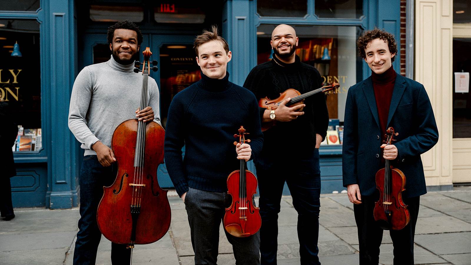Quartet artist standing in front of a blue shopfront. Each artist is holding their string instrument and smiling at the camera.