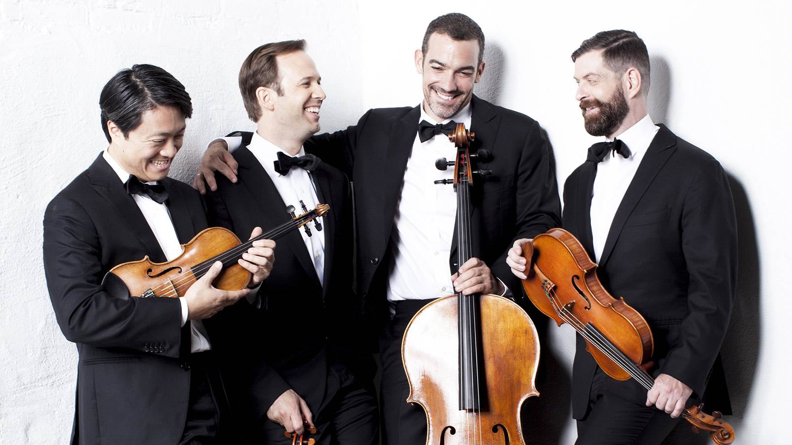 Artists of quartet posing in front of white background. They are all holding their string instruments and wearing tuxedos.