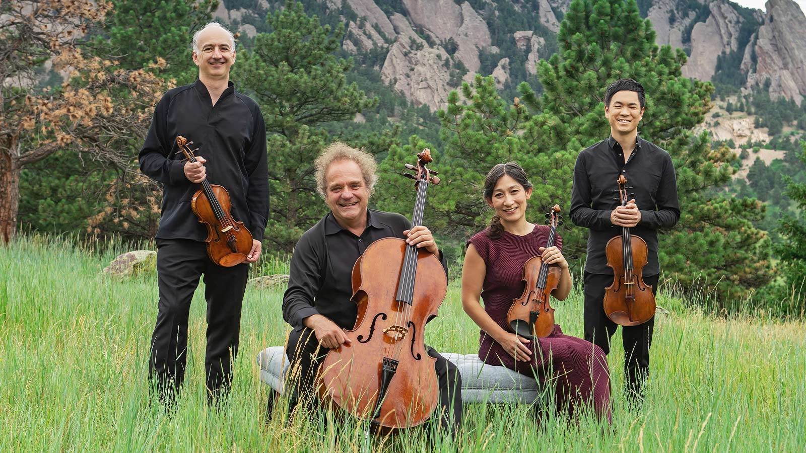Artist of quartet posing with their string instruments. They are standing in a grass area in front of a mountain.