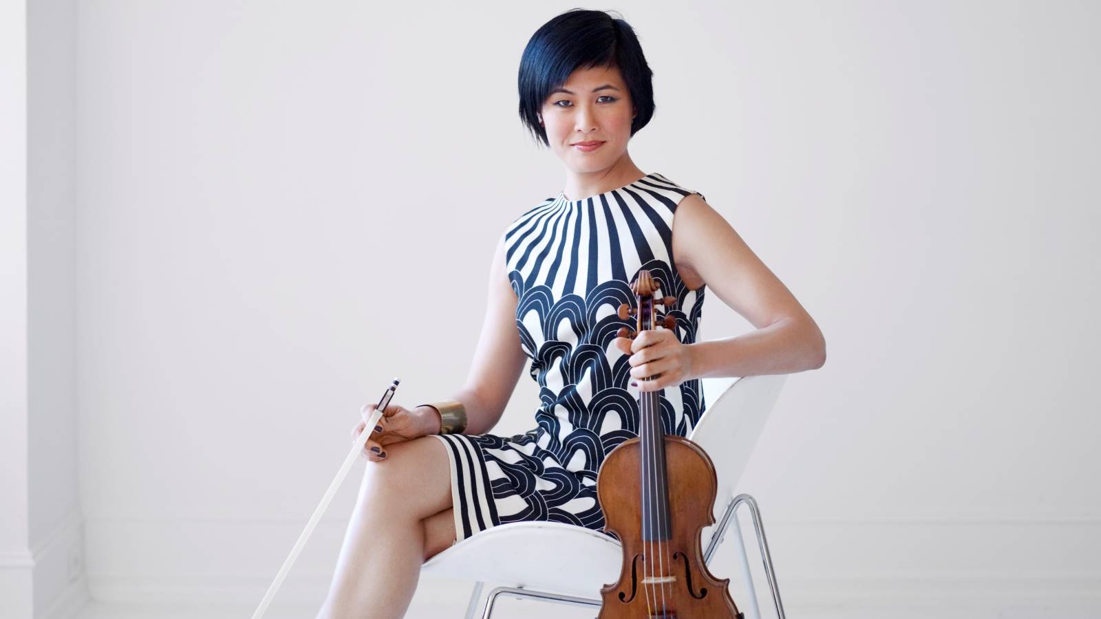 Jennifer Koh on a chair in black and white dress holding her violin and bow