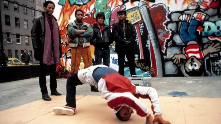 Three men watch a dancer against a graffiti wall. 