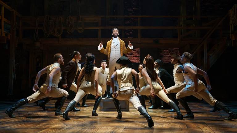 Hamilton cast performing on stage facing Alexander Hamilton