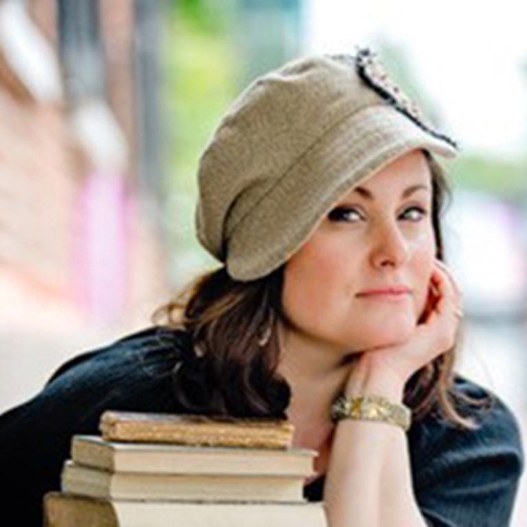 Mary Rand Hess in her hat, holding a stack of books, she gives and all-knowing smirk