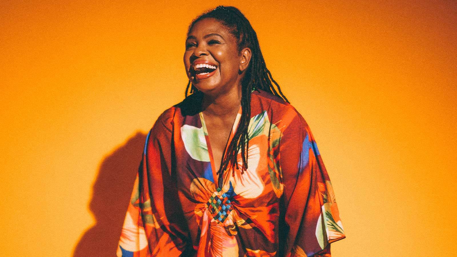 Artist Ruthie Foster against a warm yellow background. She is wearing a colorful floral dress. Her hair is in braids that are put up in a half ponytail. She is smiling toward the left.