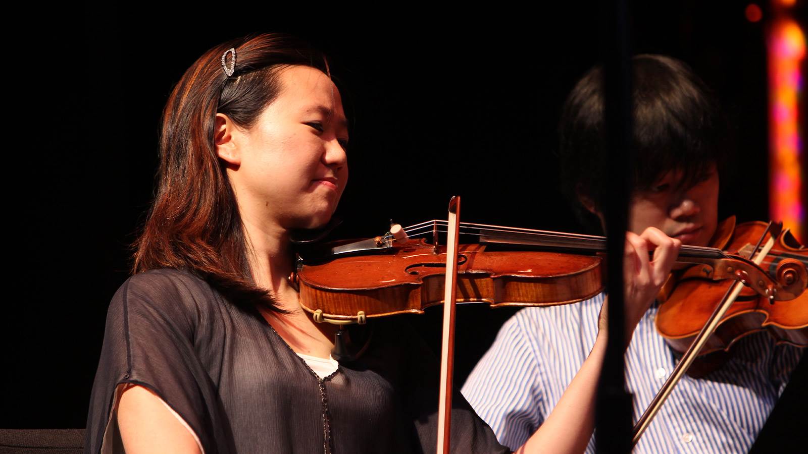 A violinist is playing her instrument. She has brown hair clipped back with a barrette in front. She is wearing a black top and is looking to the right. There is another violinist is behind her who is wearing a stripped button down. 