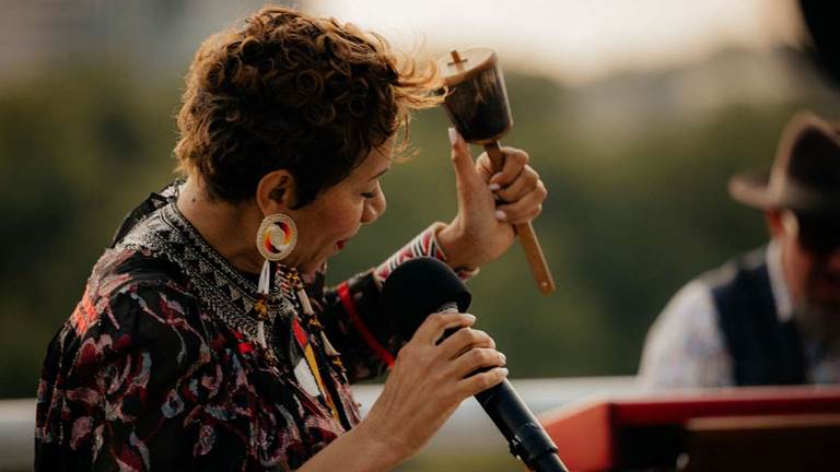 Woman with large earrings and colorful shirt sings into a microphone while holding a shaker