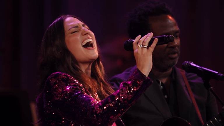 Sara Bareilles closes her eyes as she sings into a microphone with head tiled back.
