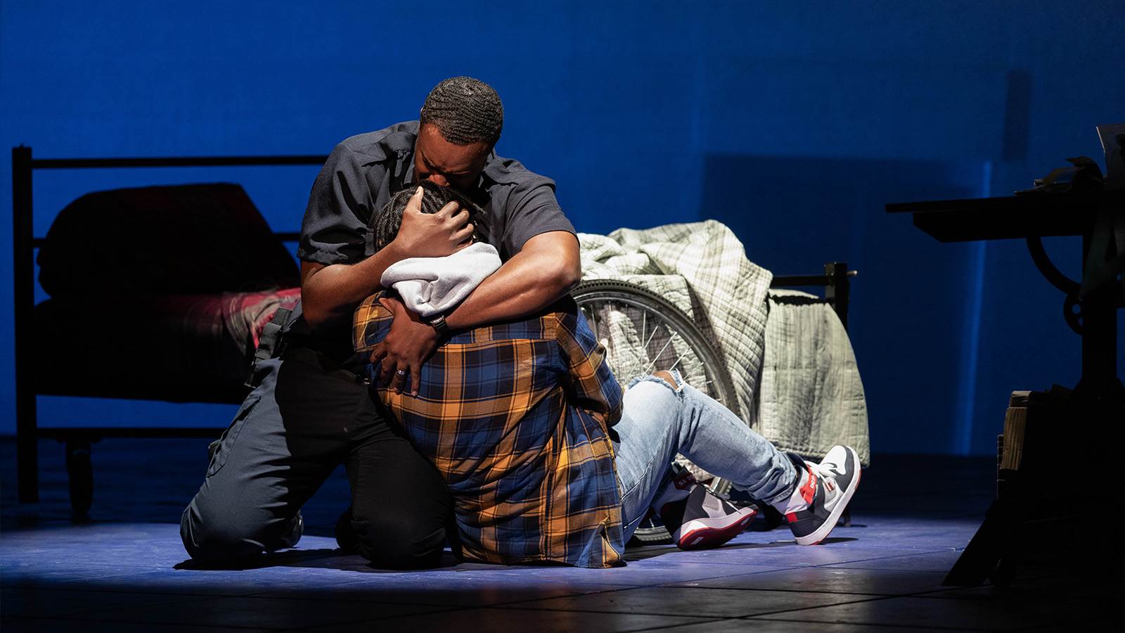 A black man kneels on the floor holding his teenage son close to his chest