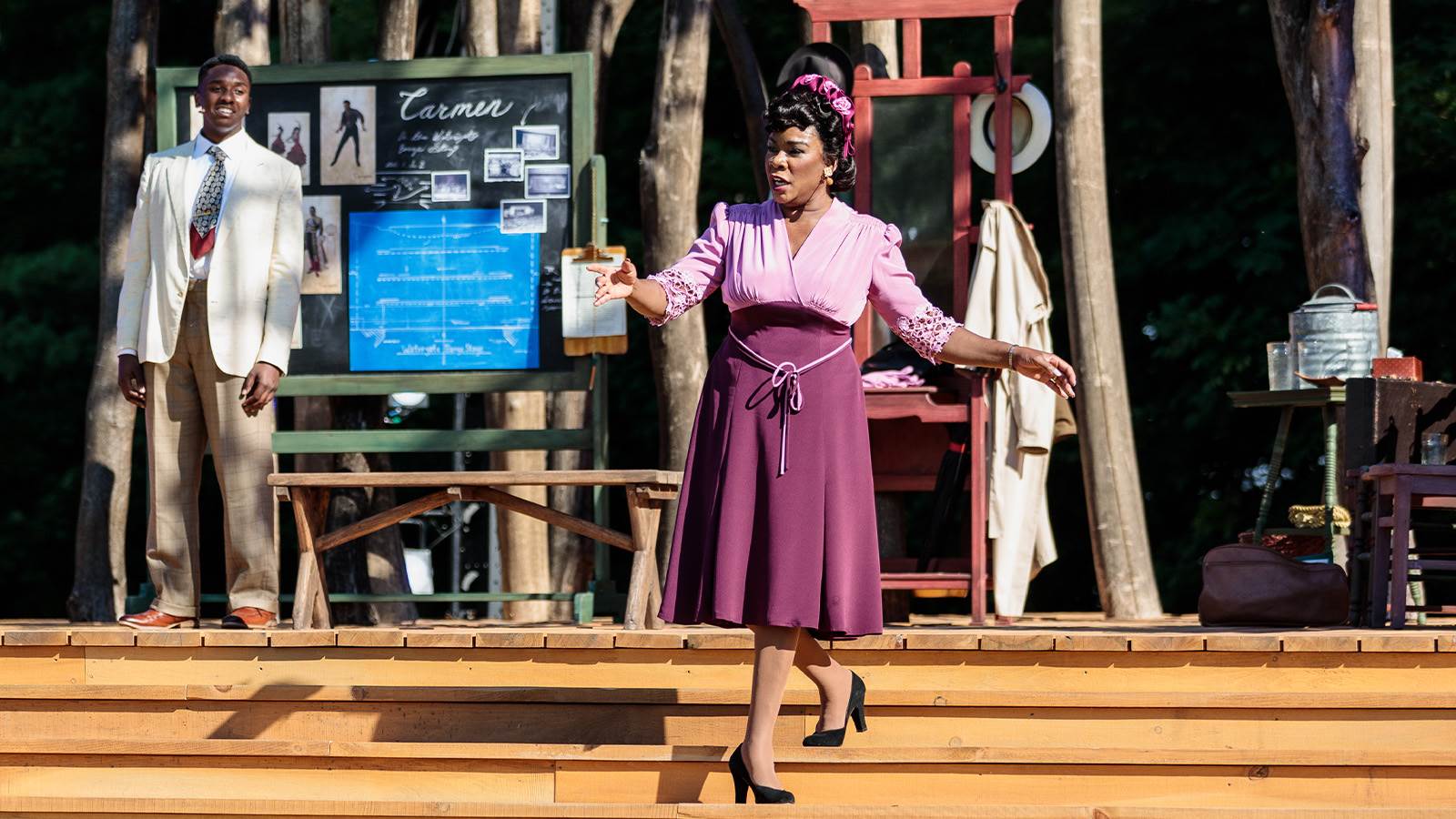 A black woman in a 1950s style purple dress walks down a staircase while singing